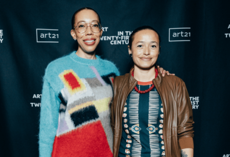 Amy-Sherald and Rose-B.-Simpson posing for a photo in front of a Art21 backdrop. on in a multicolor sweatshirt and glasses and the other in a black top with tan blue and red detail, red necklace and brown jacket