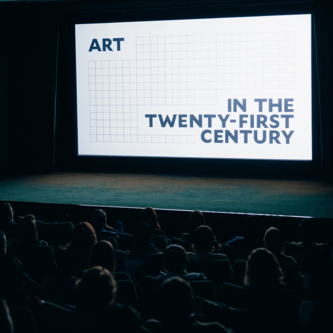 Audience sitting in a dark theater with "Art in the Twenty-First Century" displayed on the screen.