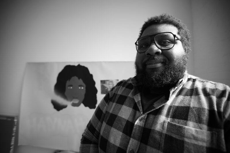 David Legget, head shot photo in black and white with flannel shirt with poster in background of a black character