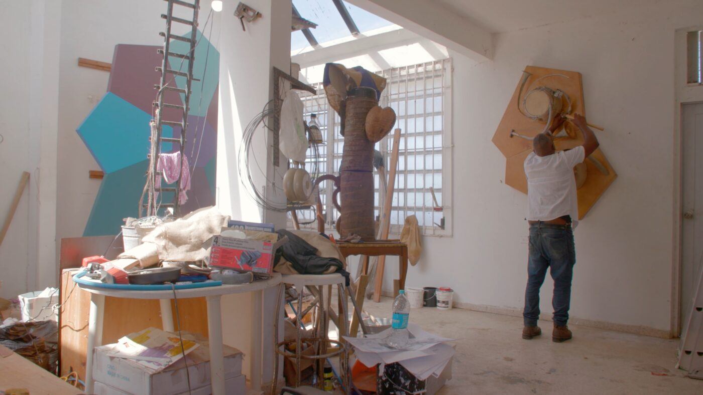 Daniel Lind-Ramos with arms raised and hammer in hand, assembling a sculpture on the wall of his studio.