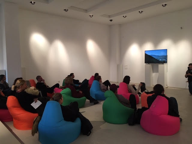 People sitting in colorful bean bag chairs on the floor of a gallery watching a screen on the far wall.