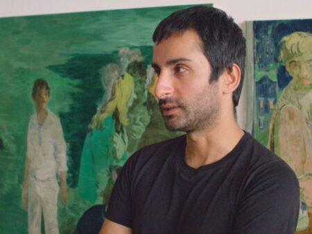 Salman Toor in his studio looking off in the distance, behind him are two large scale paintings with primarily green palettes.