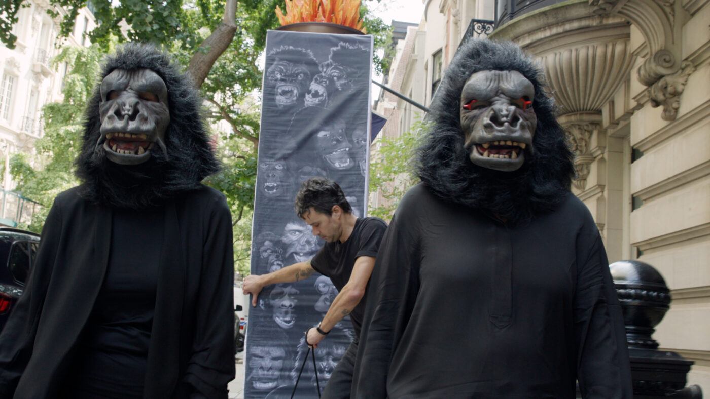 Two people wearing gorilla masks and black clothing walking down a street, behind them is a large display depicting multiple gorilla faces.