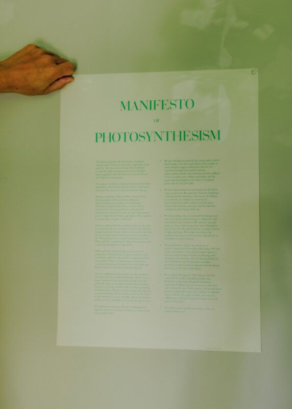 A hand holds a white sheet of paper against a surface, on the sheet are words printed in green ink, legible at the top of the sheet it reads "Manifesto of Photosynthesism."