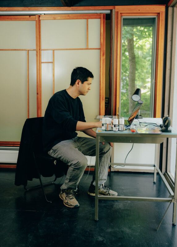 Michael Wang is in profile, seated at a desk indoors in his studio, in front of him is a laptop which he is staring at and working on.