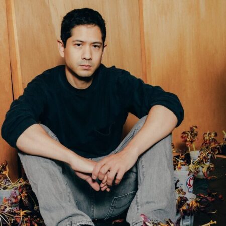 Michael Wang sitting on the floor of his studio, surrounding him are pots of wilted, purple tulips, with dead leaves scattered on the dark floor.