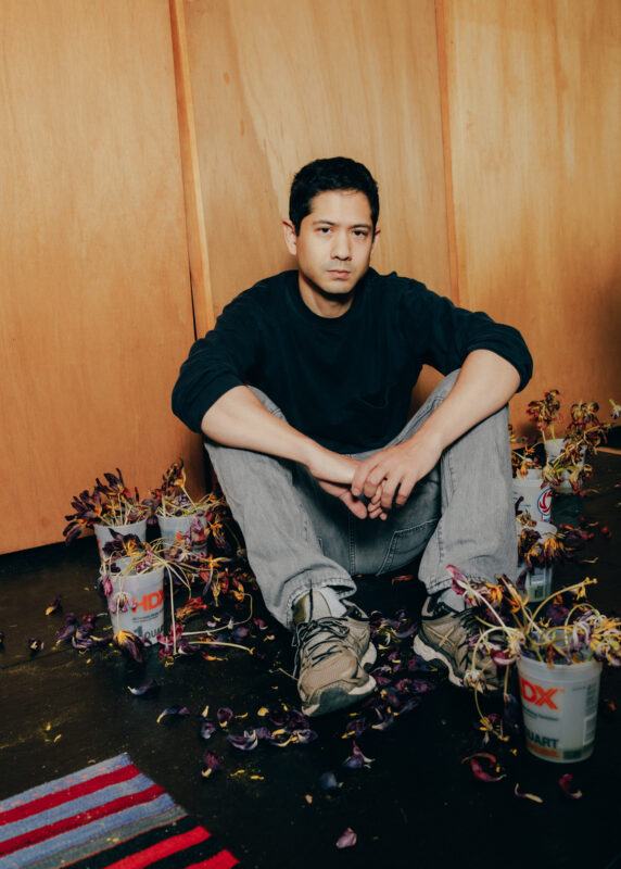 Michael Wang sitting on the floor of his studio, surrounding him are pots of wilted, purple tulips, with dead leaves scattered on the dark floor.