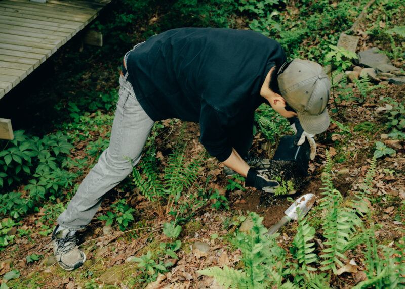 Michael Wang, outside in a wooded area, bending over to scoop a small plant into a black pot with his hands.