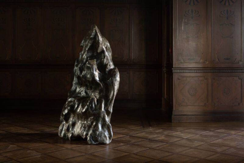 One sculpture resembling a stone is installed on the floor, the walls and floor of the room are a dark wood.