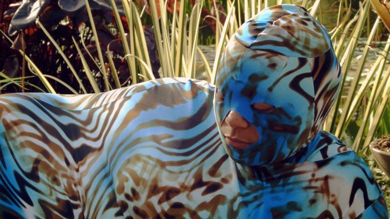 Person wearing a black and blue patterned fabric over their body, laying down in tall grass.
