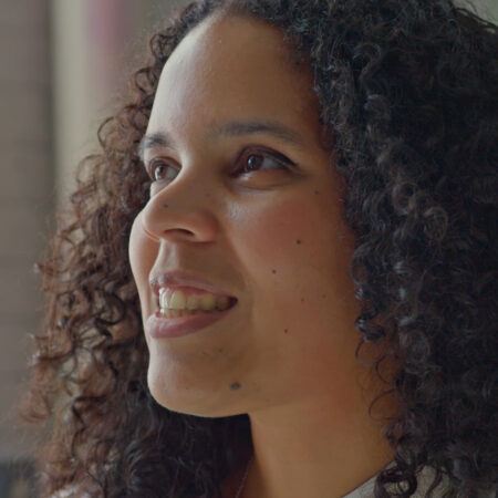 Joiri Minaya with curly hair smiling and looking to the side in a softly lit indoor setting.