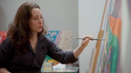 Aliza Nisenbaum holding up a paintbrush to a colorful canvas.