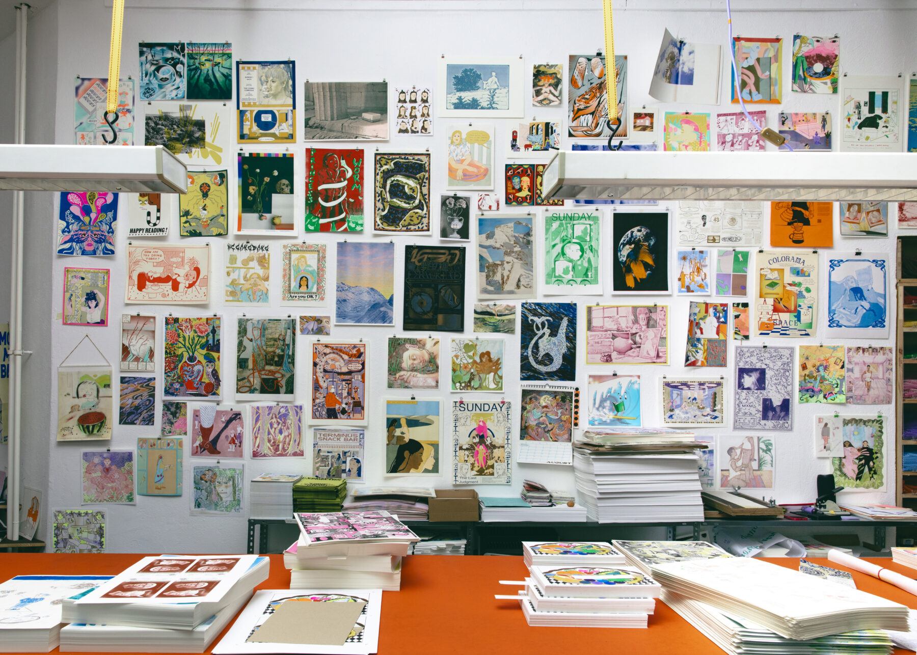 Colorama studio walls with illustrated images tacked all over. An orange table with stacks of printed books is in front.