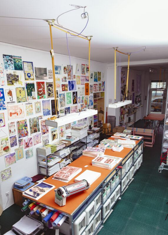 Colorama project space, with images and prints hanging on the wall from floor to ceiling and an orange table in the middle of the room with book materials on it.