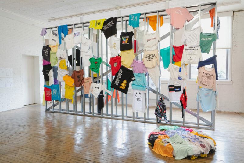 Installation of multicolored t-shirts hanging off of a large metal frame, in a gallery.