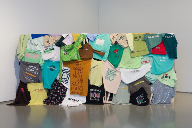 Installation of multicolored t-shirts arranged on a constructed wall in a large white room.