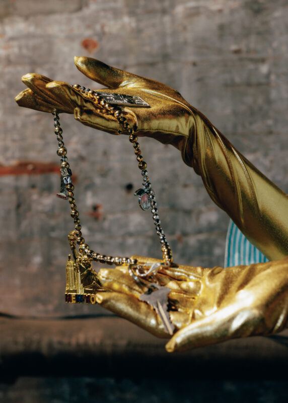 Close up of gold gloved hands holding a chunky necklace with charms and keys hanging off of it.