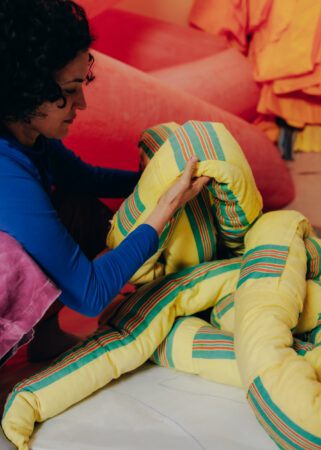 Tamar Ettun sitting in front of a colorful installation, looking down at a yellow inflatable sculpture in her hands.