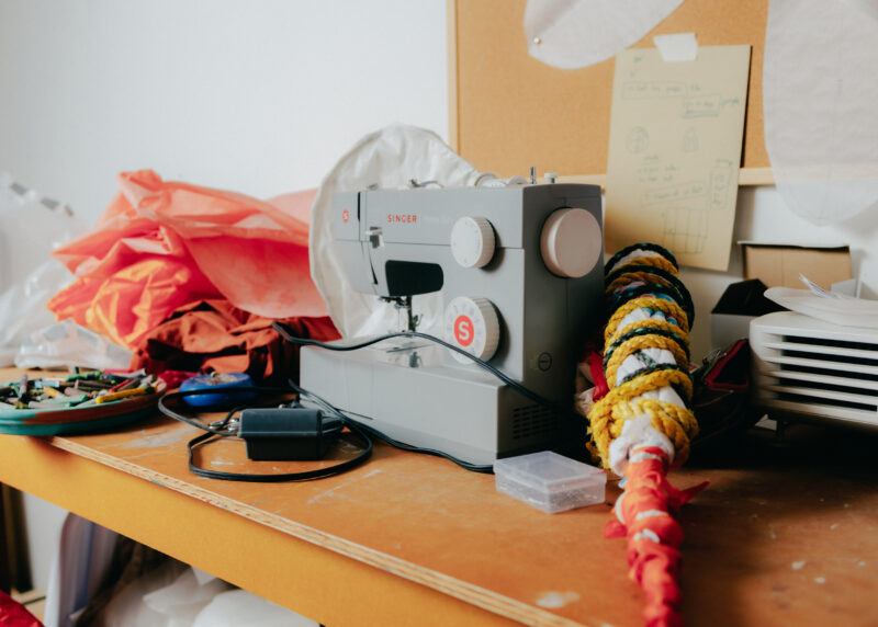 A table in the studio with a grey sewing machine on it and red fabric to the left of the machine.