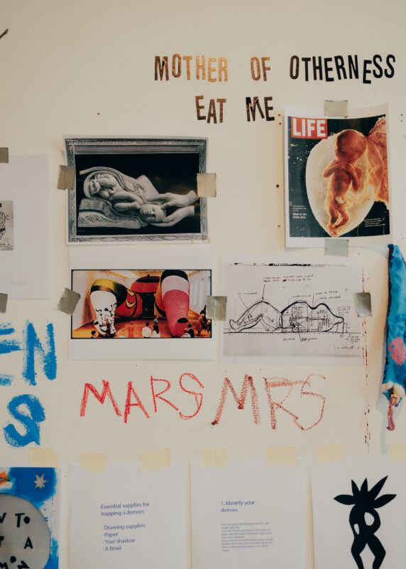 A wall in Tamar Ettun's studio with images and papers taped to the wall, above them are the words "MOTHER OF OTHERNESS EAT ME."
