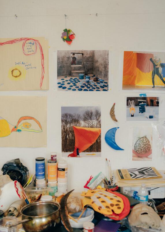 A table in the studio full of art materials, above it hangs images tacked to a white wall.