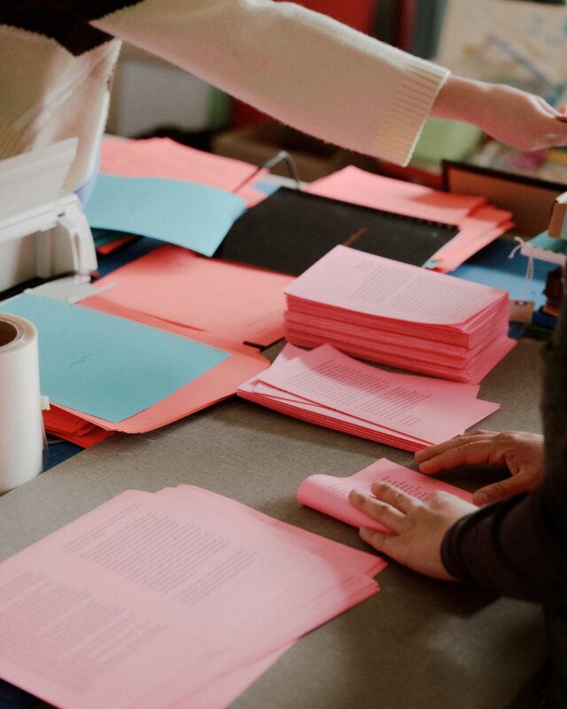 Hands assembling pamphlets of bright pink, blue, and coral papers.