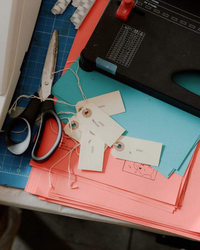 Close up of a studio table with scissors, paper ID tags, and blue and pink printed papers.