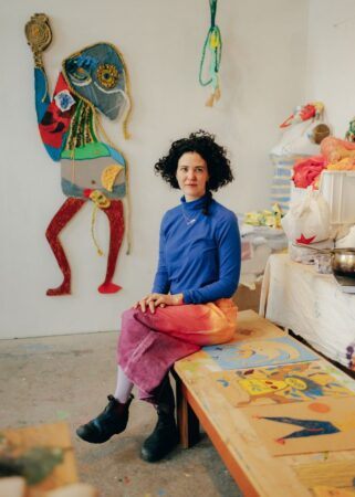 Tamar Ettun seated on a wooden bench in her studio with artwork hanging on the wall behind her.
