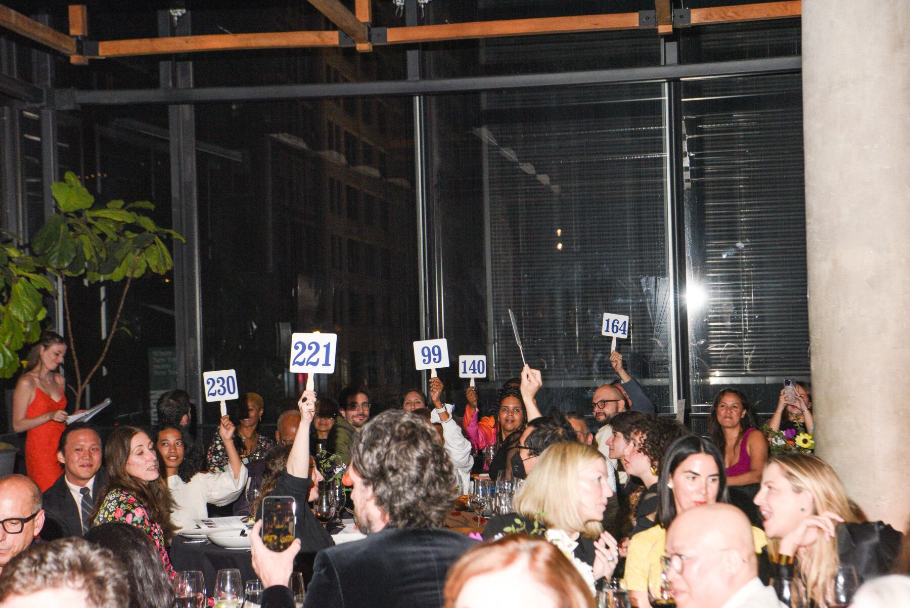 Room of people seated at dinner tables with bidding paddles raised above their heads.