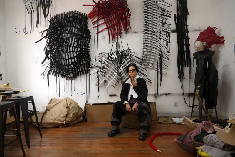 Bárbara Sánchez-Kane sits on a box in her studio. The space is cluttered with various materials and artworks.