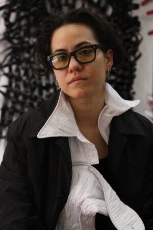 Bárbara Sánchez-Kane with short dark hair and glasses, wearing a black coat and a white, high-collared inner garment, looks into the camera against a blurred dark and white background.