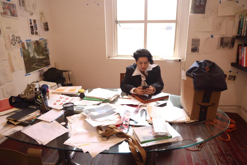 Bárbara Sánchez-Kane sitting at a cluttered desk in a sunlit room, looking at a phone. The desk is surrounded by papers, books, and other items, with a window in the background.