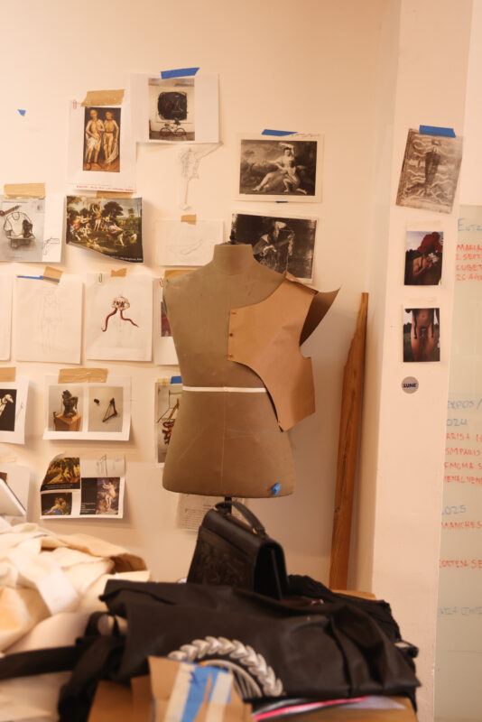 Mannequin with a paper garment in progress stands in a workspace filled with sketches and fabric pieces.