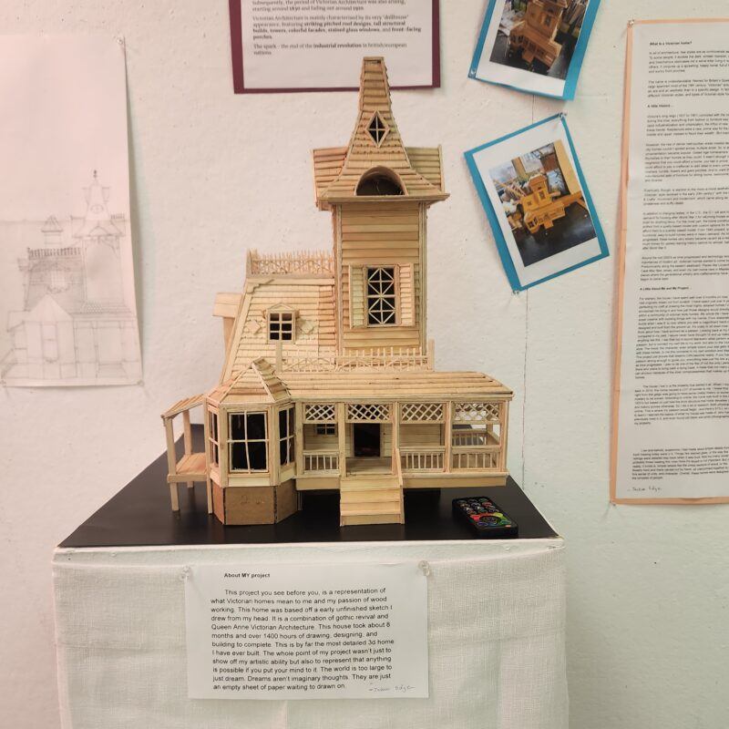 A detailed wooden model of a house displayed on a black stand, accompanied by descriptive text and illustrations on the wall behind it.