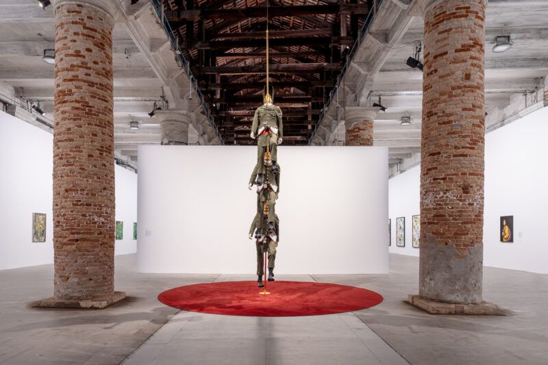 Sculpture of three identical figures stacked vertically, placed on a red circular carpet in an art gallery with brick pillars and multiple artworks hung on the walls.