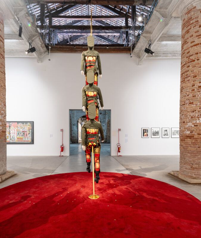 Sculpture of three identical figures stacked vertically, placed on a red circular carpet in an art gallery with brick pillars and multiple artworks hung on the walls.