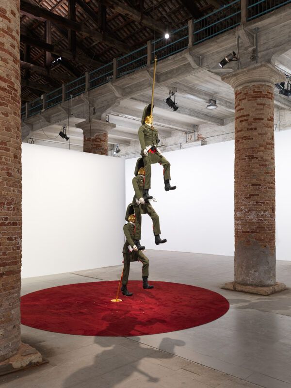 Sculpture of three identical figures stacked vertically, placed on a red circular carpet in an art gallery with brick pillars and multiple artworks hung on the walls.