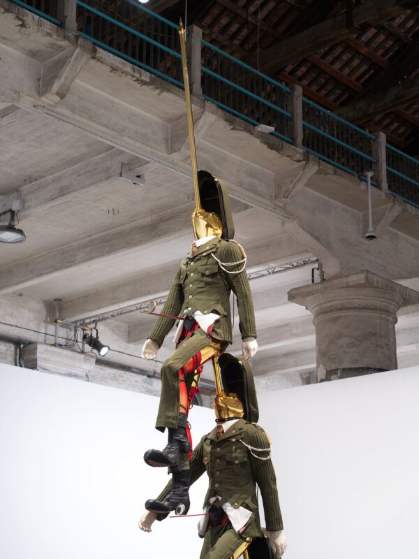 Two mannequins in military uniforms are suspended vertically from the ceiling by poles, which go through their bodies.