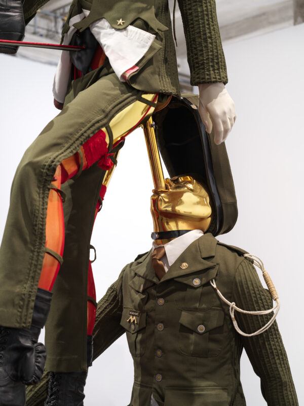 Two mannequins dressed in military-style uniforms positioned in an unusual, artistic arrangement with a pole connecting the one on top to the one below through its mouth.