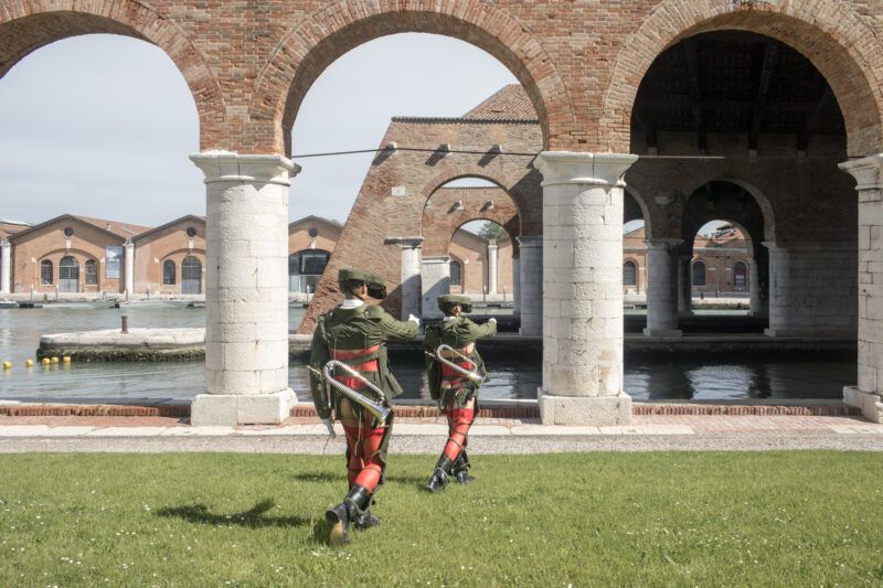 Two performers dressed as soldiers in uniform walk under brick arches by a waterfront. The background features historical brick buildings and calm water.