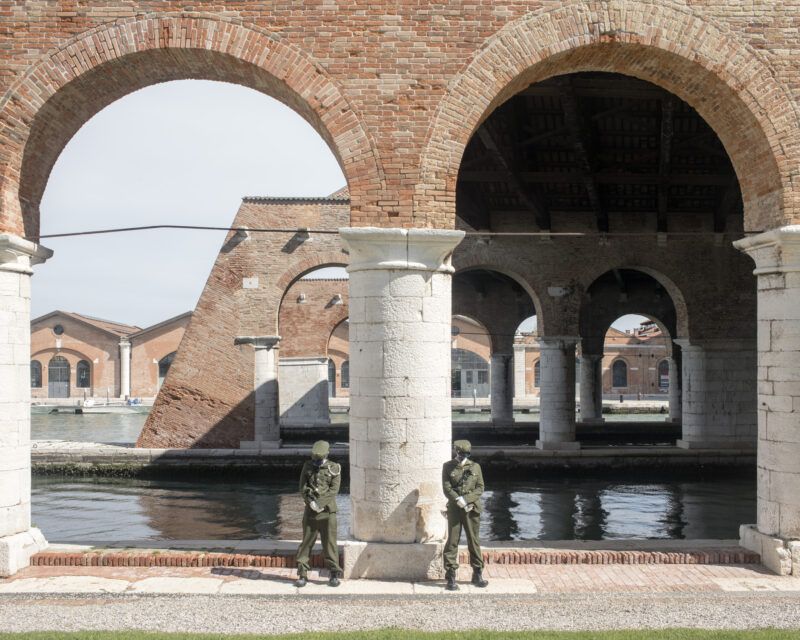 Two performers dressed as soldiers stand near a brick and stone archway, with water and additional brick structures visible in the background.