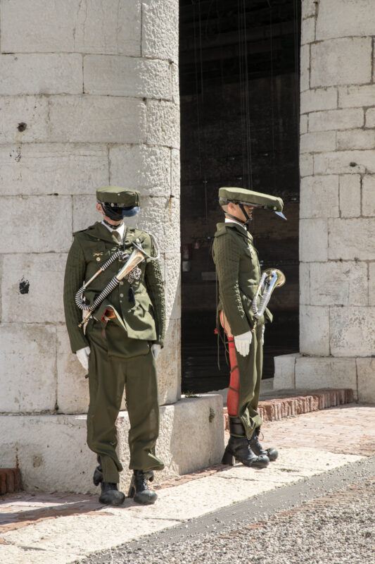 Two performers dressed as uniformed guards stand near a white stone wall, each holding a musical instrument. One faces forward and the other faces sideways. They wear green attire, hats with elongated brims, and gloves.