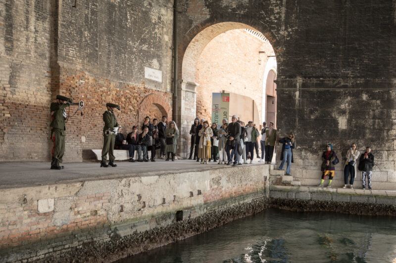 A group of tourists observe two performers dressed as uniformed guards performing a demonstration under a large archway in a brick structure by a canal.