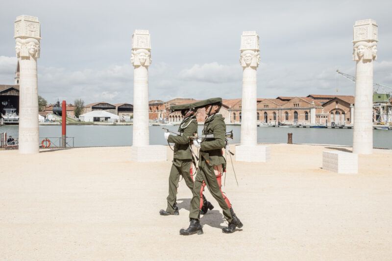 Two performers dressed as uniformed soldiers walk holding rifles in front of four white columns, with a waterfront and industrial buildings in the background.