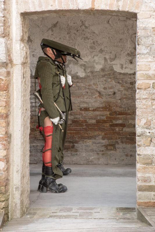 Two performers dressed in antiquated green military uniforms stand in a brick and stone archway, carrying long swords and wearing hats with long brims.