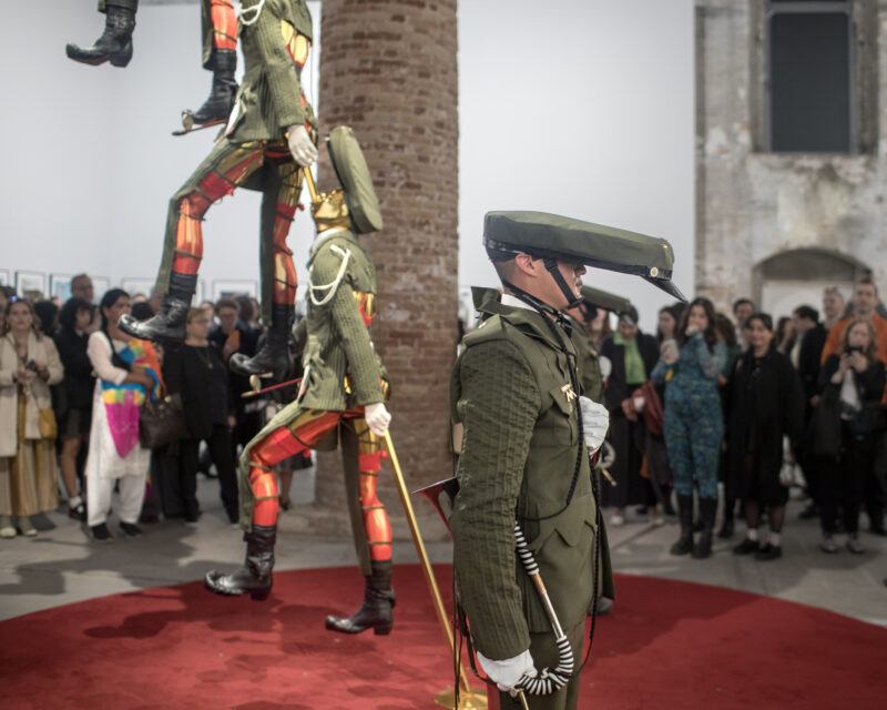 A costumed performer dressed in a green military-style outfit with an elongated hat stands in front of a crowd, while two mannequins in matching outfits are suspended in mid-air.