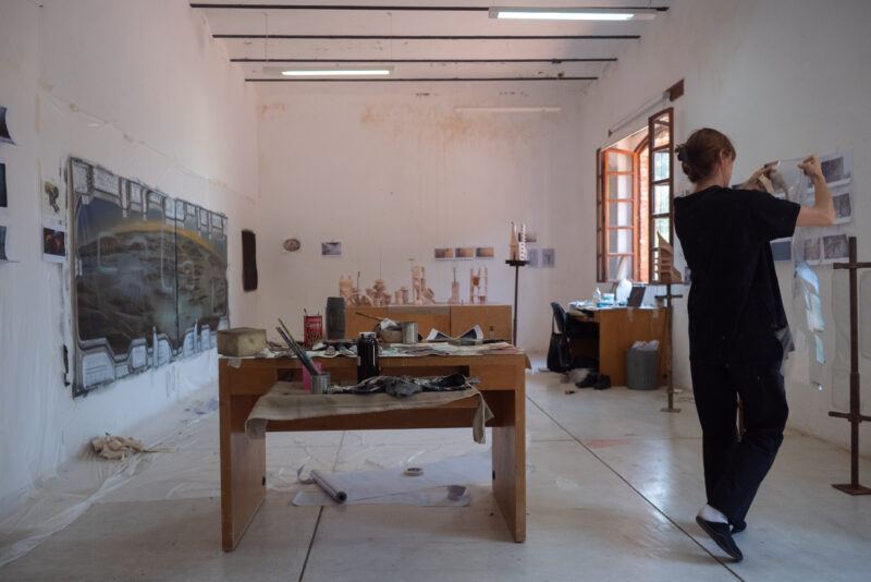 Carolina Fusilier with her back turned holding up a sheet of clear plastic in her stuido, a wooden table covered in art supplies is in the center of the room.