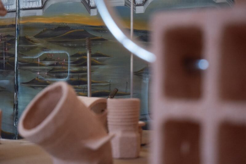 Out of focus image of clay sculptures in the foreground and a painted landscape with hills and antennas in the background.