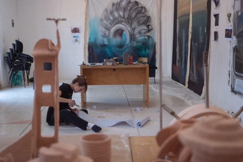 Carolina Fusilier sitting on the floor of her studio, surrounded by sculptures and art supplies, holding scissors to cut a large sheet of paper.