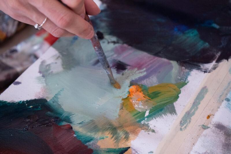 A hand holding a paintbrush to a palette, blending green and orange.